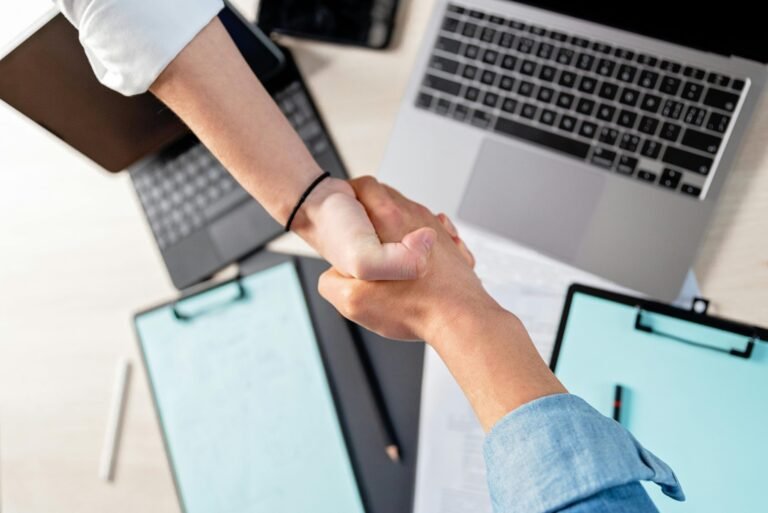 Two people shaking hands over laptops and documents in an office, symbolizing a business deal.